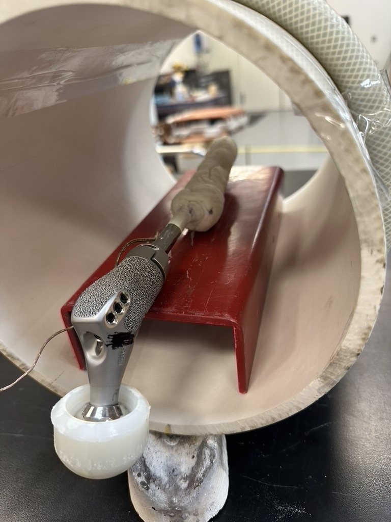 A cemented hip stem resting on a red bench fixture inside a large plastic tube, with a thermocouple wire attached to the implant.